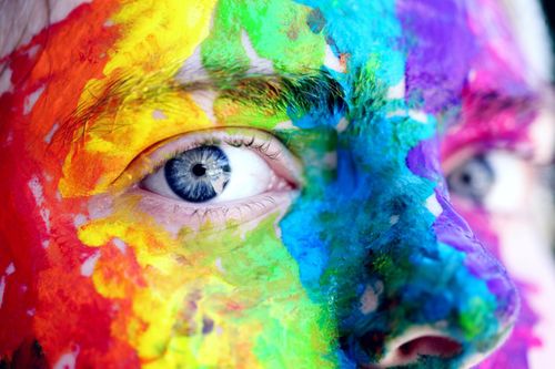 A creative with blue eyes stares ahead with colorful paint covering her face.