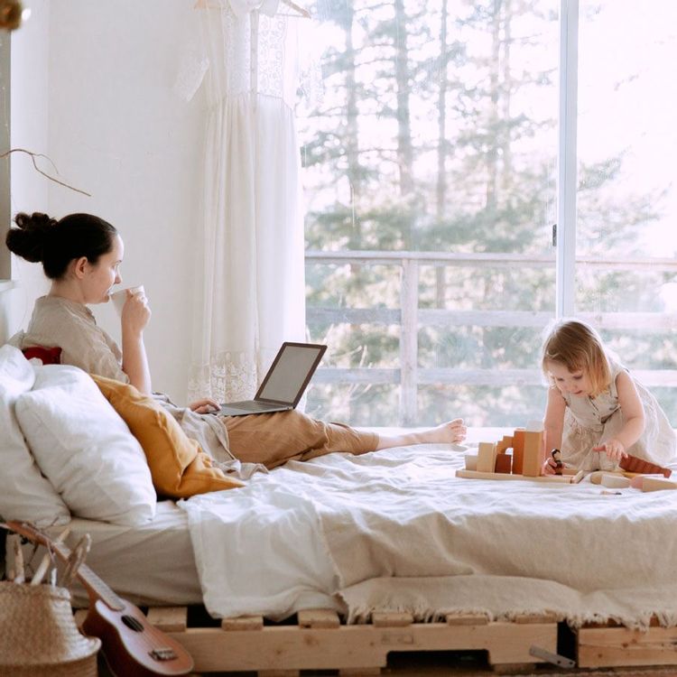 Mom sitting on bed with coffee and laptop with toddler playing on bed