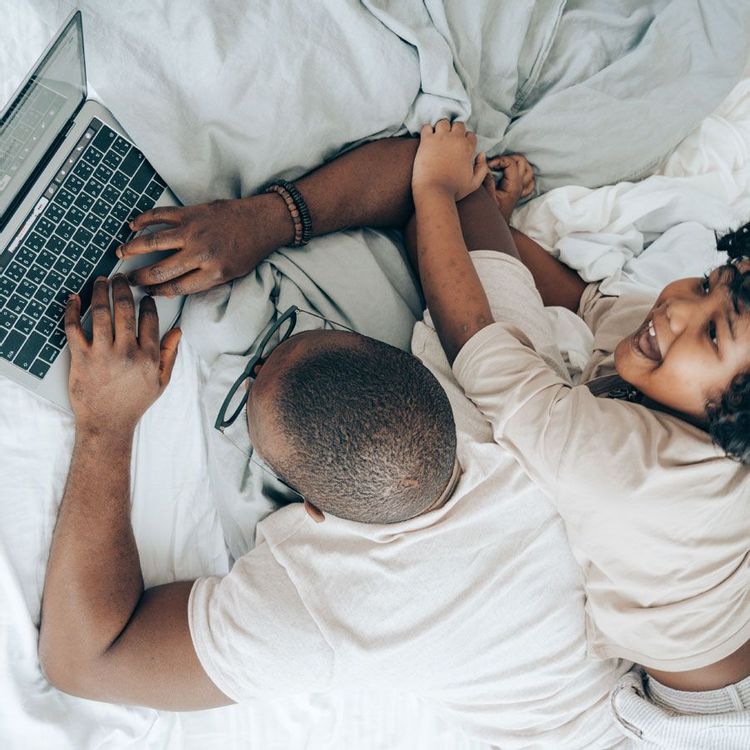 Man laying on bed working on laptop while his daughter players with him