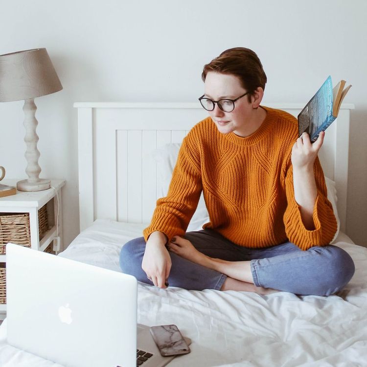 Woman on a bed with a book and a laptop.